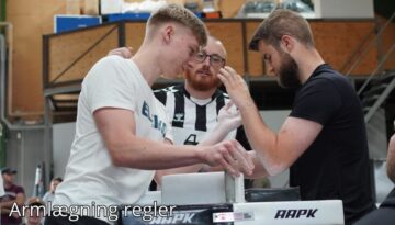 Billedet viser to mænd, der gør sig klar til en armlægningskamp ved et professionelt bord. En dommer i sort-hvid-stribet trøje overvåger dem nøje for at sikre, at de følger reglerne. Baggrunden viser en indendørs hal med tilskuere og andre deltagere, der skaber en konkurrencestemning. Billedetekst: "Armlægningsregler''